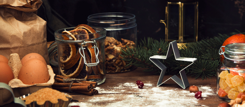 Świąteczny stół do wykrawania pierniczków – widać mąkę, foremki, jajka, gałązkę świerku, cynamon, jabłka i cukier, tworzące przytulny, bożonarodzeniowy klimat.